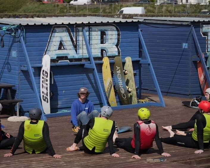 A group of people in wetsuits sitting on a wooden deck, with surfboards leaning against a blue building. 4