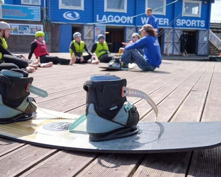 A group of people sitting on a wooden deck with surfboards and wetsuits, in front of a building with 'LAGOON WATER SPORTS' signage. 1
