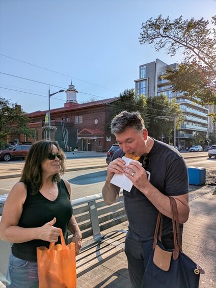 Two people standing on a sidewalk, one eating a sandwich and the other holding a shopping bag. 2