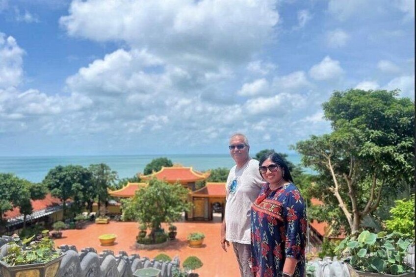 A couple standing on a terrace overlooking a traditional building with a red roof, surrounded by greenery and a clear sky. 5
