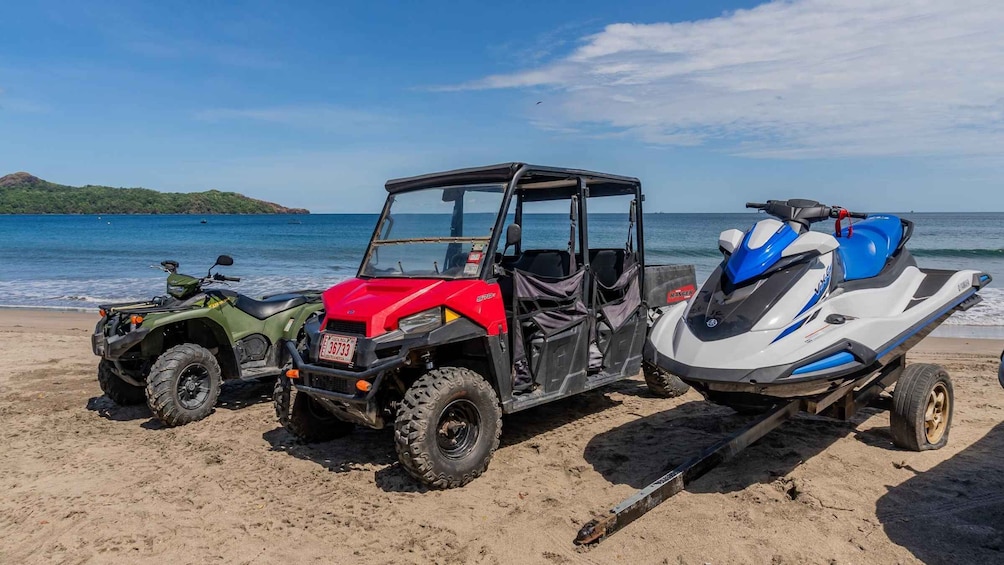 A red ATV, a green ATV, and a blue and white jet ski on a sandy beach with a clear blue sea and a distant island in the background. 5