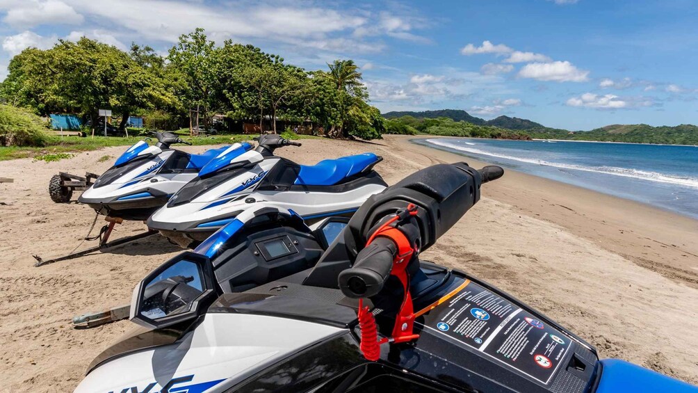 Jet skis lined up on a sandy beach with a clear view of the ocean and a forested hill in the background. 2