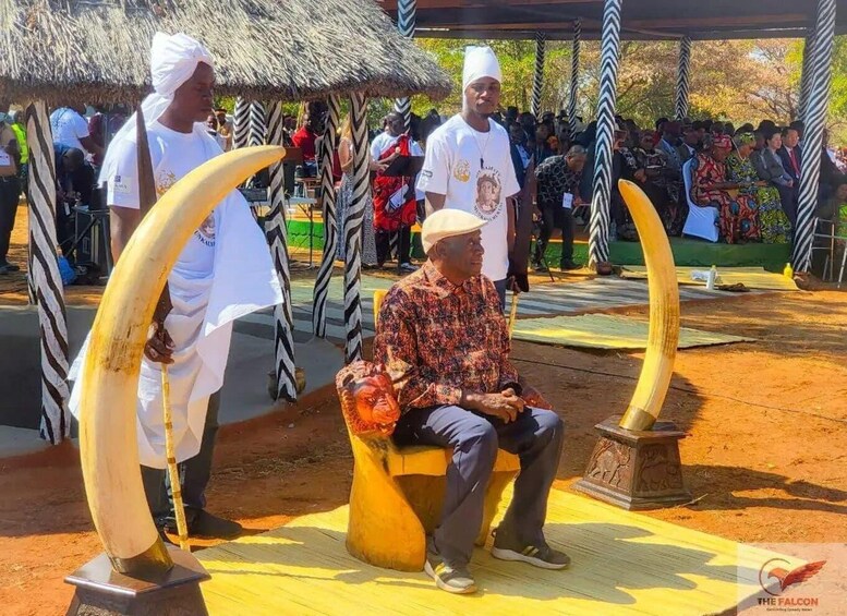 A man seated on a bench under a thatched roof structure, with large tusks displayed on pedestals in the foreground. 2