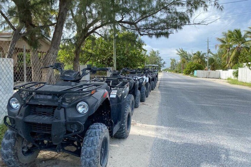 A row of ATVs parked on a road with trees and a fence in the background. 3