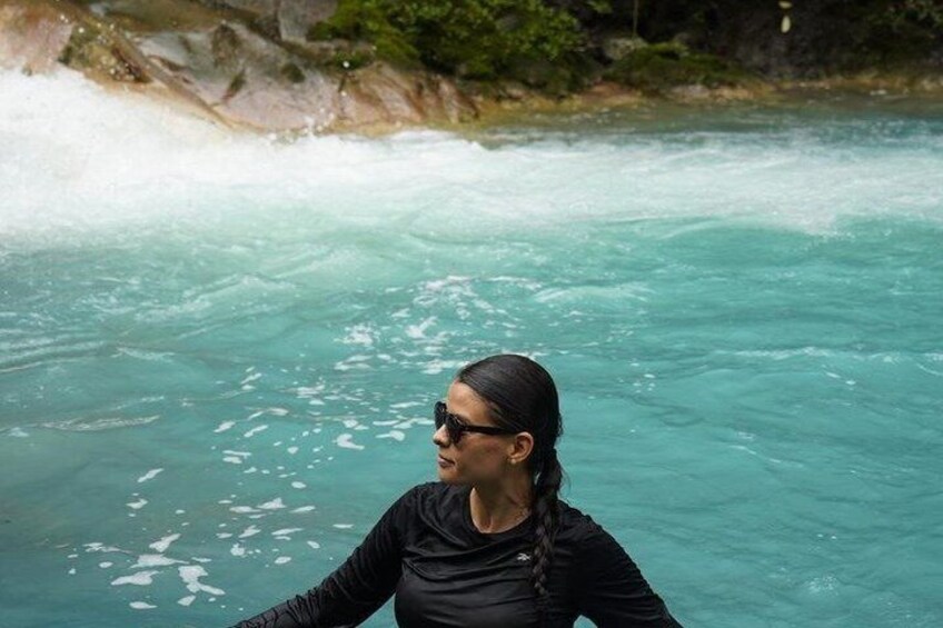A person in a black top and long pants, standing in clear turquoise water with a rocky shoreline in the background. 3