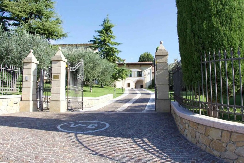 A cobblestone pathway leading to a gated entrance with a VEB logo, surrounded by neatly trimmed hedges and trees. 2