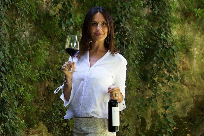 A person holding a wine glass and a bottle of wine, standing in front of a leafy background. 4