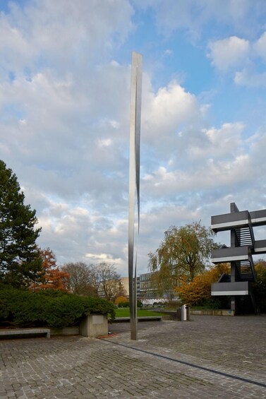 A tall, slender metal sculpture in an open plaza with cobblestone ground, surrounded by trees and modern buildings. 2