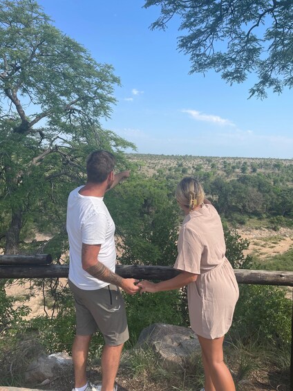 Two people holding hands, overlooking a savanna landscape with scattered trees and a clear sky. 2