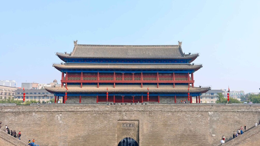 A traditional multi-tiered building with red pillars and blue accents, situated in front of a stone wall with an archway. 2