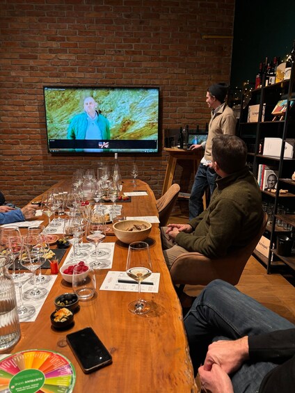 A group of people seated at a long wooden table with wine glasses and a bowl of fruit, in a room with brick walls and a television screen. 2