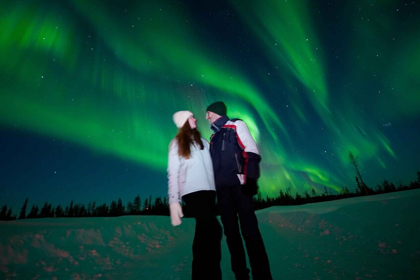 Two people standing on a snow-covered field under a night sky with green aurora borealis. 4