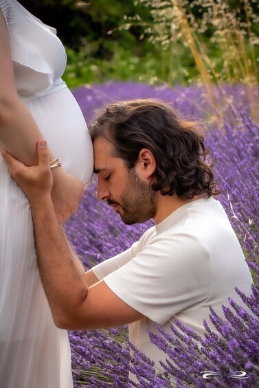 A pregnant woman in a white dress holding her belly, surrounded by purple flowers. 2