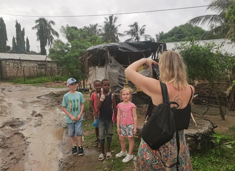 A group of children and an adult in a muddy outdoor area with a makeshift shelter and palm trees in the background. 3