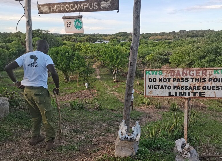 A man in a white shirt with 'CAMP GEDEVG' on the back, standing in a vineyard with a sign warning of a KWS danger area and a Vietato Passare Quest limit. 5