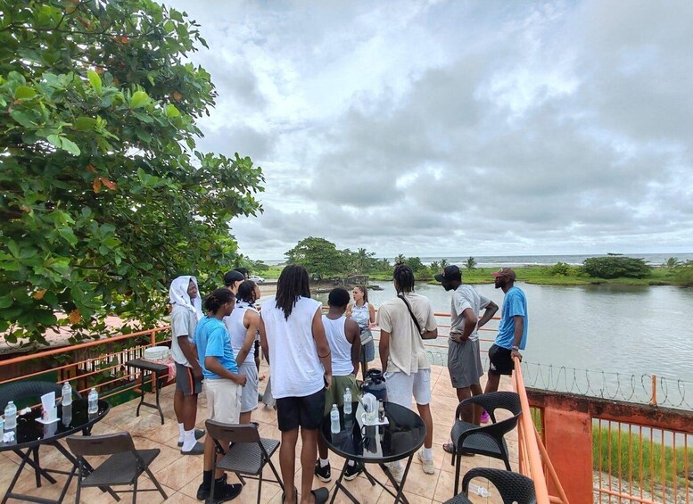 A group of people standing on a terrace overlooking a body of water, with trees and a cloudy sky in the background. 3
