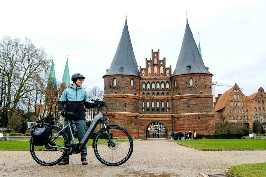 A person with a bicycle in front of a historic brick building with two pointed towers. 1