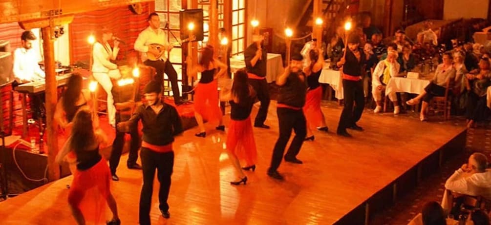 A group performing a dance on a wooden stage with an audience seated in the background. 1