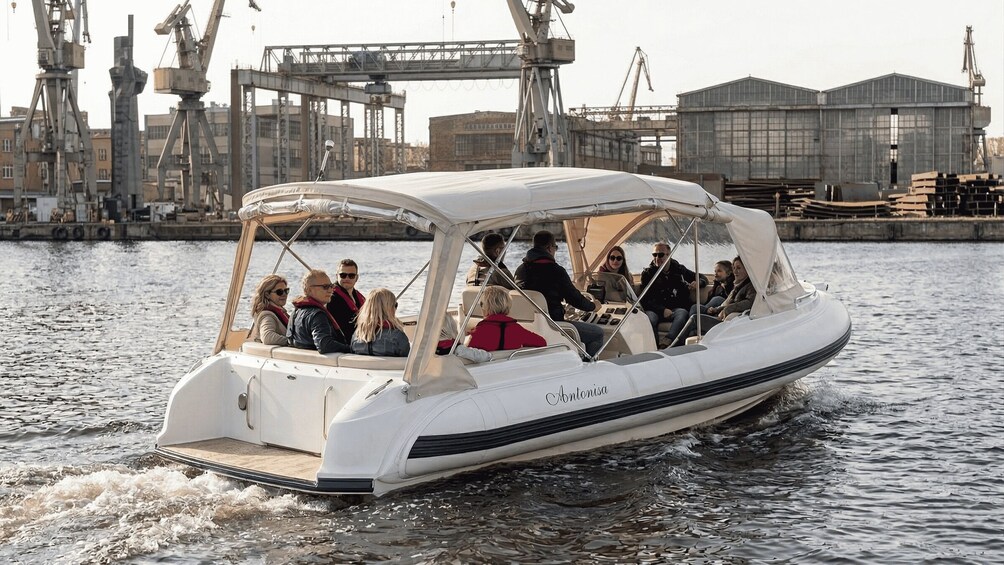 A white boat with 'Antonisa' written on the side, carrying passengers on a body of water with industrial structures in the background. 4