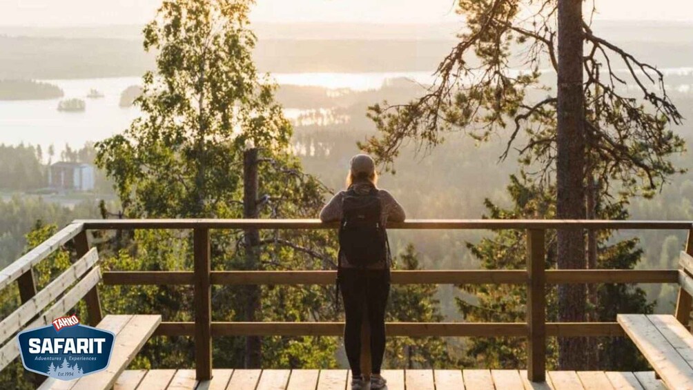 A person standing on a wooden deck overlooking a forested landscape with a body of water and buildings in the distance. 3