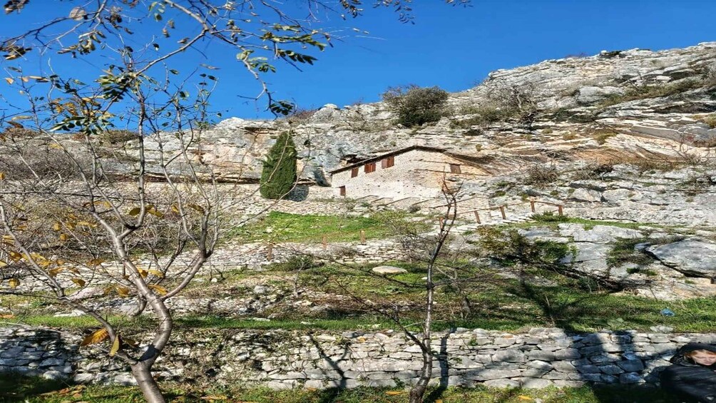 A stone building with a sloped roof, surrounded by rocky terrain and sparse vegetation. 1