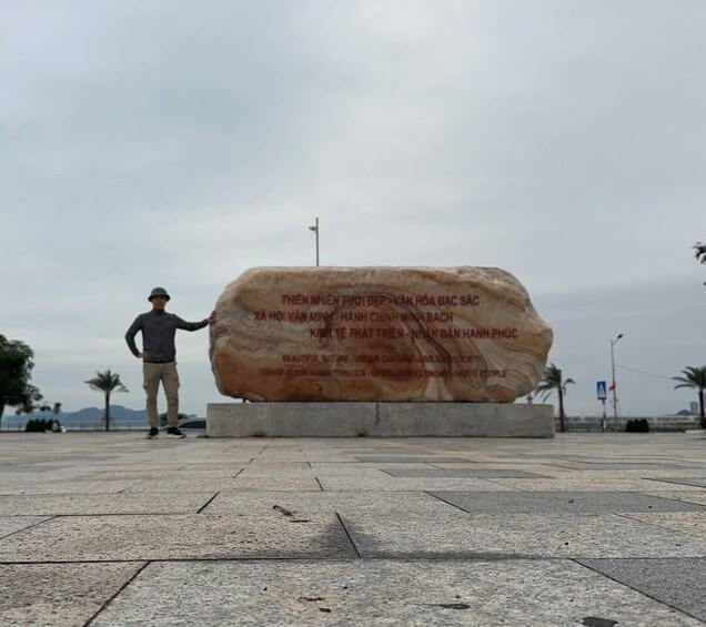A large stone monument with inscriptions, a person standing beside it, and a paved area with palm trees in the background. 3