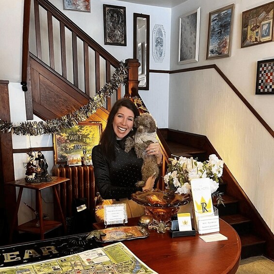 A person holding a dog in a hotel lobby with a Christmas tree, framed artwork, and a sign reading 'QUIET TIME 11:00 PM 7:00 AM'. 4