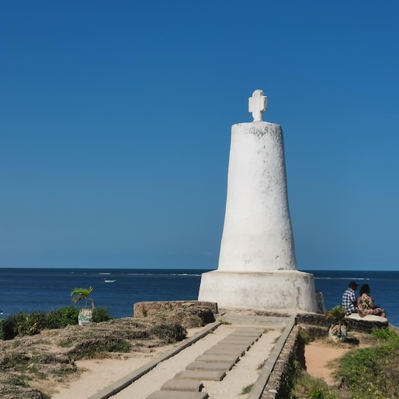 A white monument with a figure on top, located by the sea with a clear sky. 1