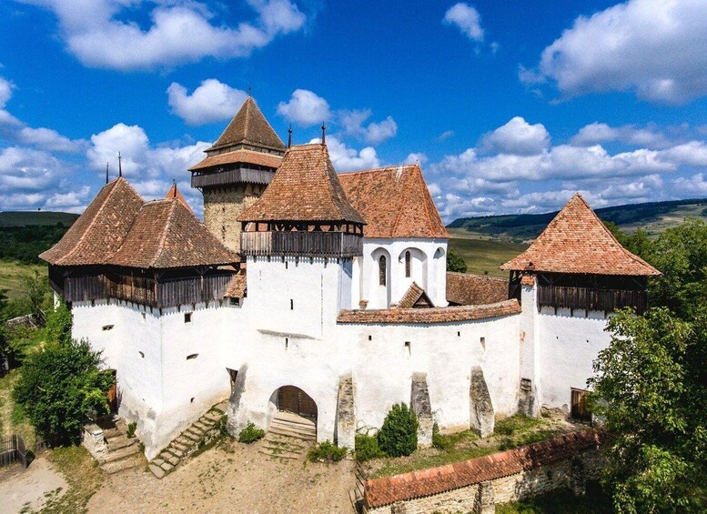 A medieval castle with white walls and red-tiled roofs, surrounded by greenery and hills. 1