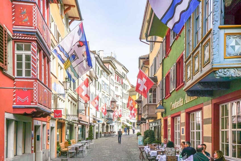 A cobblestone street lined with colorful buildings, flags, and outdoor seating areas. 3
