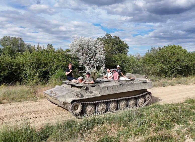 A group of people on a military tank in a natural setting with trees and a dirt path. 3
