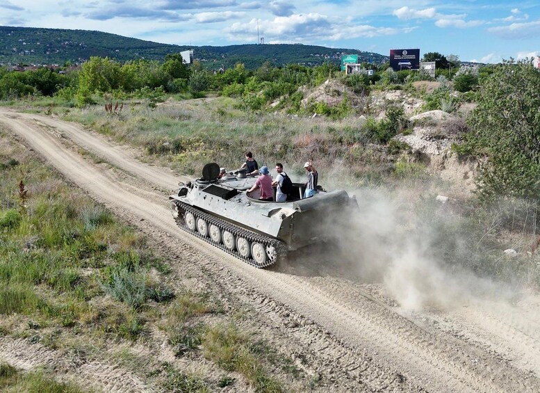 A military tank moving on a dirt road with people on top, surrounded by greenery and hills in the background. 5