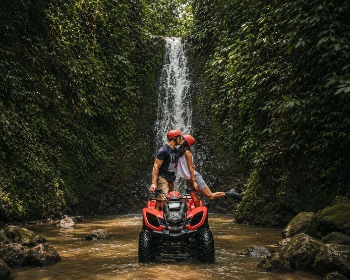 A person riding a red ATV through a forested area with a waterfall in the background. 2