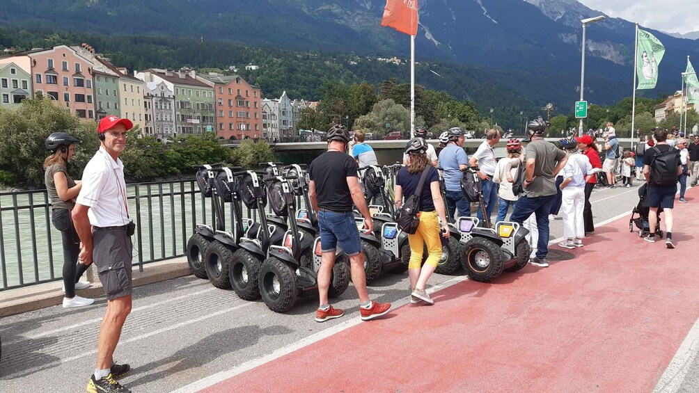 A group of people standing on a bridge with Segways lined up, overlooking a town with colorful buildings and mountains in the background. 3