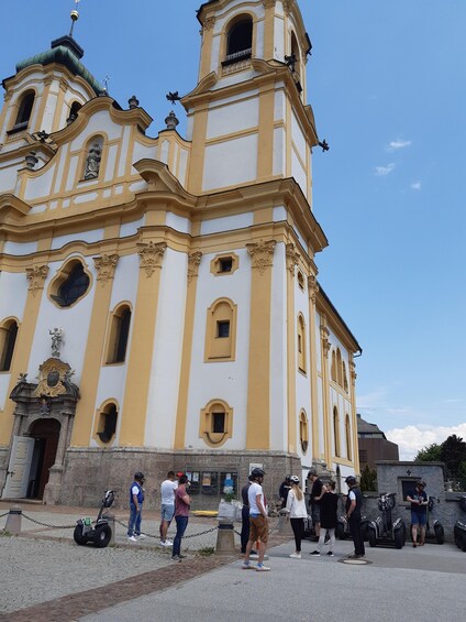 A historic church with yellow and white facade, a bell tower, and statues. People are standing in front, some on scooters. 2