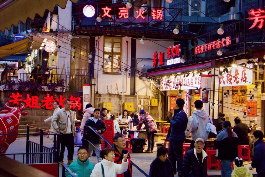 A bustling street scene with people gathered around food stalls, traditional architecture, and neon signs. 1