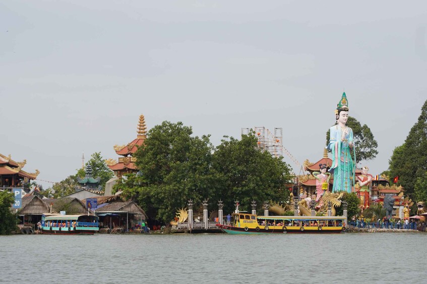 A large statue of a woman by a river, traditional buildings, and a boat with passengers. 1