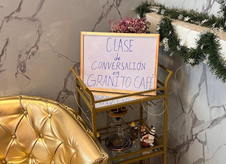 A sign reading 'CLASE de CONVERSACIÓN en GRANITO CAFE' with a gold chair and a coffee machine on a metal cart. 1