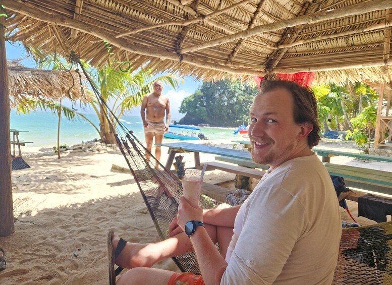 A man sitting on a beach chair under a thatched roof, holding a drink, with a beach and ocean in the background. 4