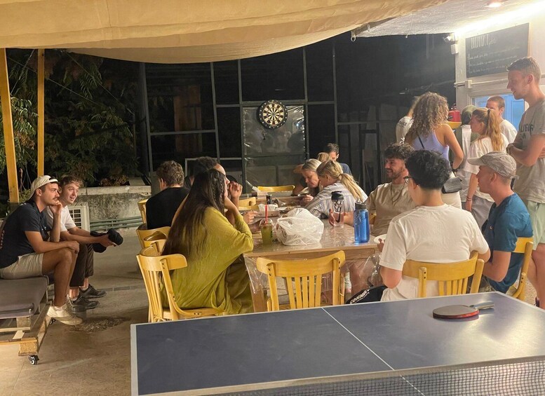 An outdoor gathering with people seated around tables, a ping pong table, and a dartboard. 1
