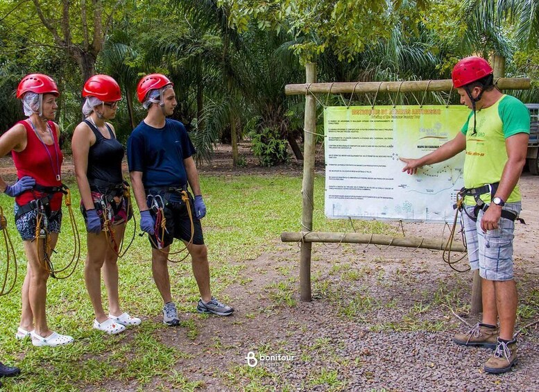 Four people in helmets and harnesses, standing in a forested area with a signboard in the background. 3