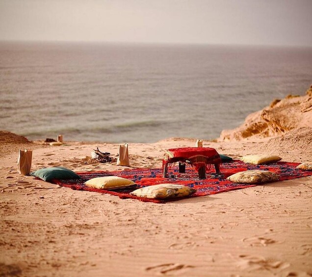 A beach with a red mat, pillows, and a small table set up on the sand. 5