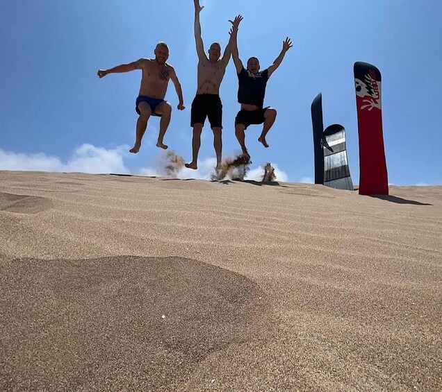 Three people jumping off a sand dune with a red and black object in the background. 2
