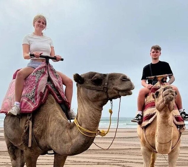 Two people riding camels on a beach. 3