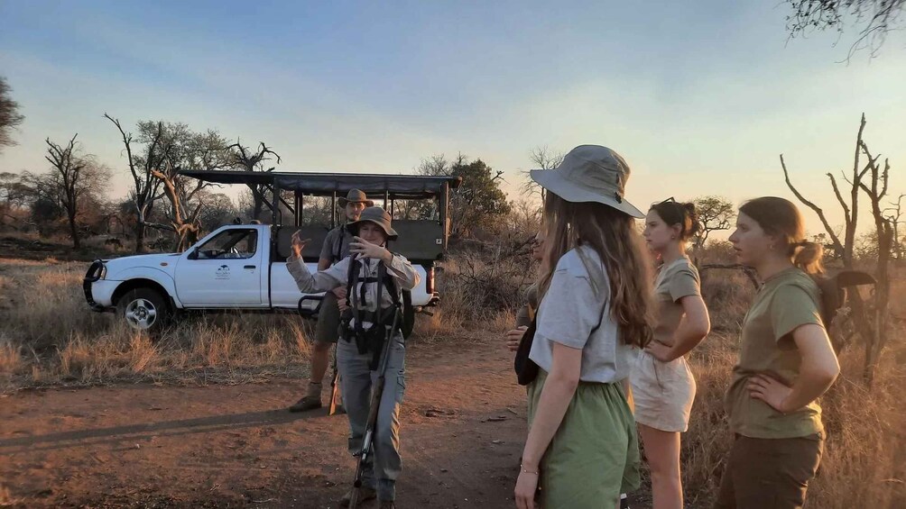 A group of people standing on a dirt path with a white pickup truck and a safari vehicle in the background. 2
