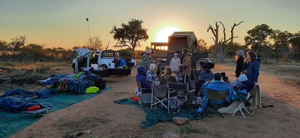 A group of people gathered around a vehicle at sunset in a desert-like setting with tents and camping gear. 4