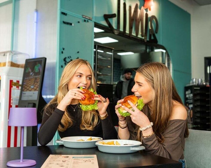 Two people eating burgers at a restaurant with a teal wall and a sign that reads 'SIMIR.' 2