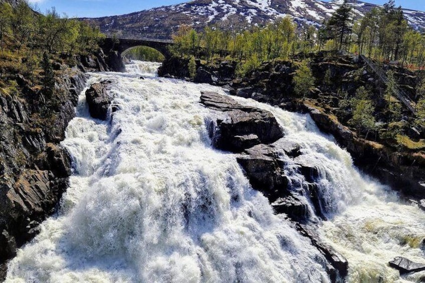 A powerful waterfall cascading over rocks, a bridge in the background, and a forested mountain range. 5