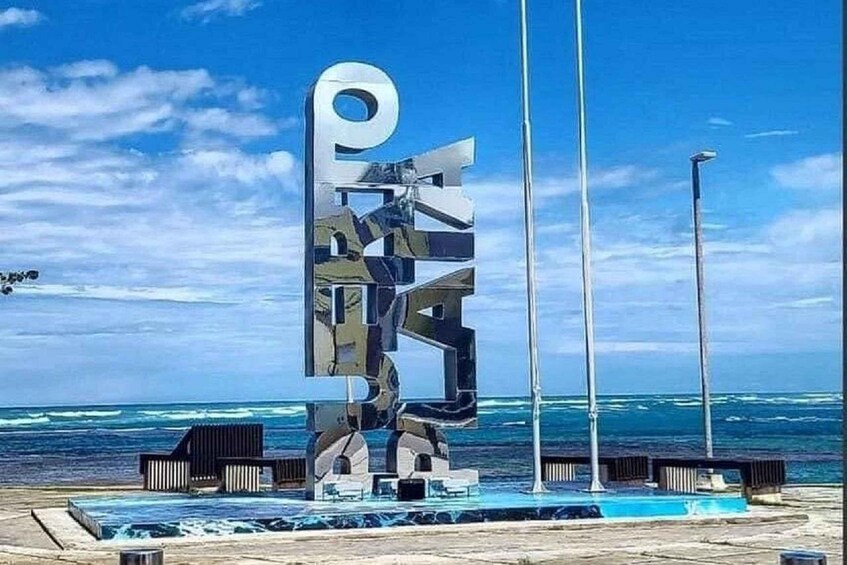 A metallic sculpture with the letters 'P' and 'E' prominently displayed, set against a backdrop of the ocean and a clear sky. 4