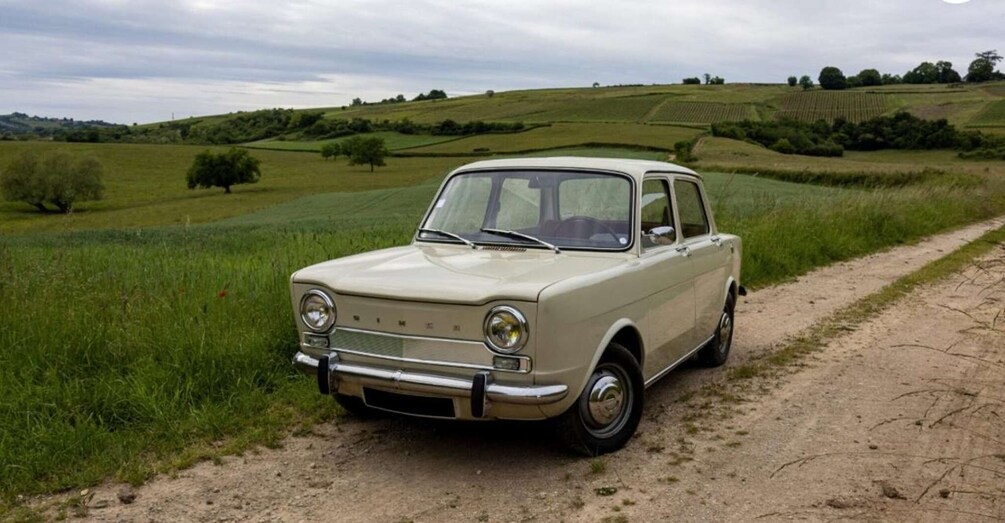 A vintage beige car parked on a dirt path in a rural landscape with rolling hills and green fields. 4